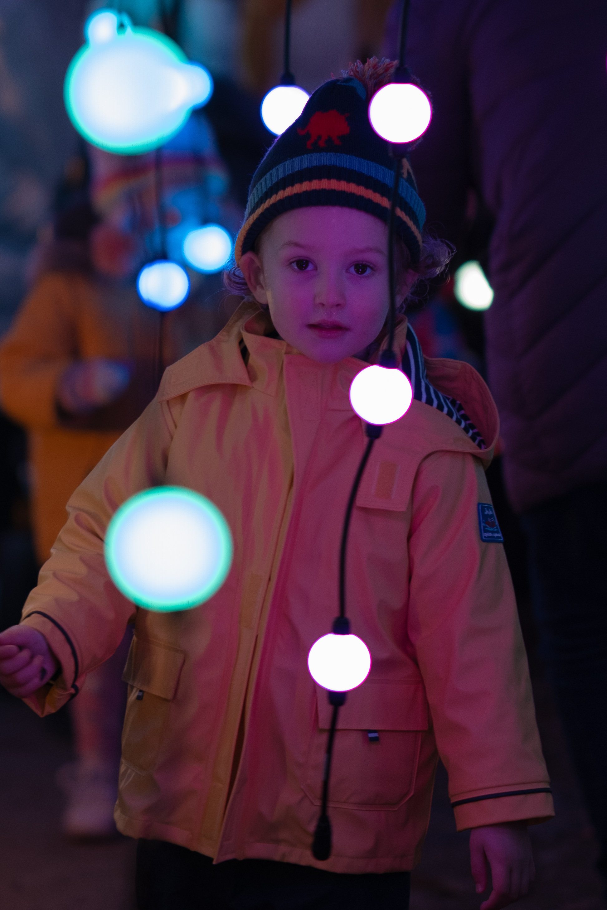 A kid amongst dangling Christmas orbs looking directly at the camera. The kid is wearing a yellow coat