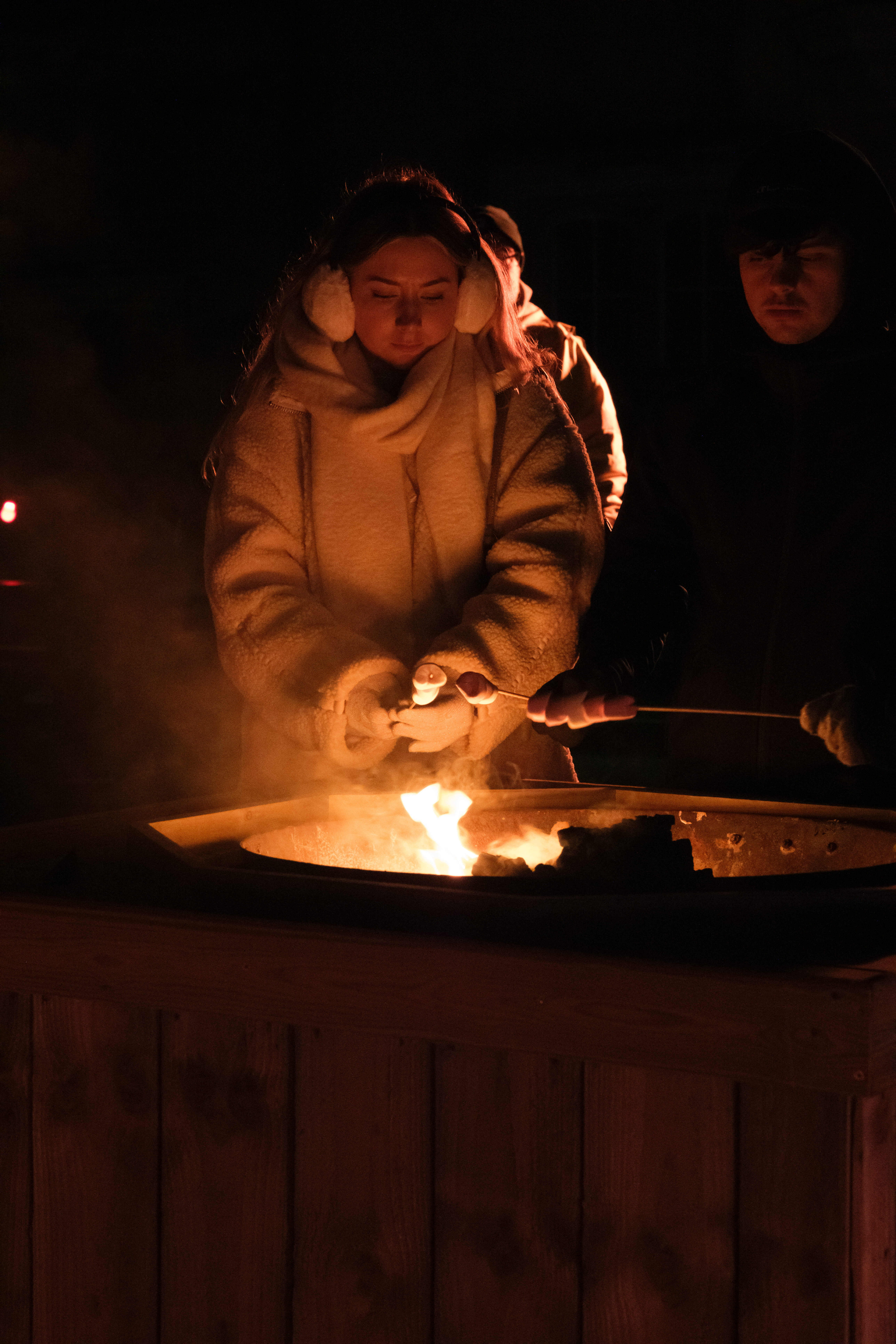 A person wearing white fluffy earmuffs and a white fluffy coat roasting marshmellows